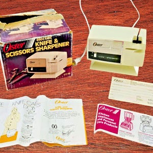 May include: An Oster electric knife and scissors sharpener, with its original box and paperwork, sits on a wooden surface. The box is purple, black, and red, with the product name and a picture of the sharpener. The sharpener is off-white.