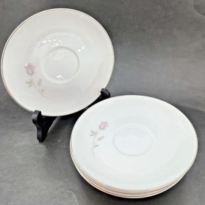 Könnte beinhalten: Weißes Porzellan-Untertassenset mit zarter rosa Rose und silberner Umrandung. Die Untertassen haben eine zentrale Vertiefung für eine Teetasse und sind gestapelt. Das Set enthält mehrere Untertassen, ideal für eine Teeparty oder ein elegantes Abendessen.
