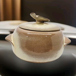 Puede incluir: Una fuente de cerámica con tapa. El plato es una gradación de colores marrón y crema. La tapa tiene un asa en forma de mariposa. El plato tiene dos asas laterales.