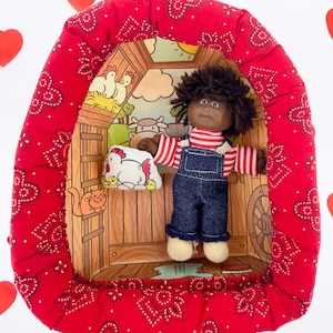 May include: A vintage doll wearing denim overalls and a striped shirt, inside a red bandana-lined frame with a farm scene backdrop. The doll holds a small chicken-shaped pillow. Red heart decorations are scattered around the frame.