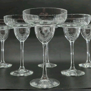 Peut inclure: Un ensemble de verres à margarita en verre transparent. Chaque verre a un large bol peu profond et une longue tige. Les verres sont disposés en rangée, les bols vers le haut. Le fond est sombre.