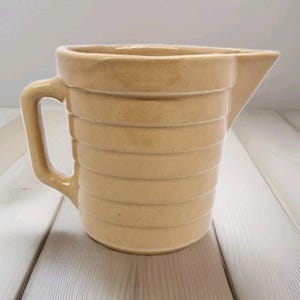 May include: A tan ceramic jug with a ribbed design. The jug has a handle and a spout.