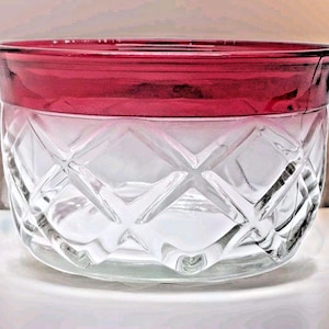 May include: A clear glass bowl with a ruby red rim. The bowl has a diamond pattern etched into the glass. The bowl is round and has a slightly flared rim. The glass is clear, allowing light to pass through.
