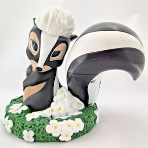 Könnte beinhalten: Eine Keramik-Stinktierfigur mit schwarz-weißem Körper, weißem, flauschigem Kopf und blauen Augen. Das Stinktier sitzt auf einem grünen Sockel mit weißen Blumen mit gelben Zentren. Die Figur hat ein cartoonartiges Design.