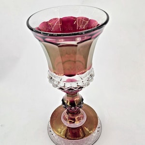 Può includere: Un calice decorativo in vetro con bordo svasato e corpo sfaccettato. Il calice presenta una ricca combinazione di colori iridescenti oro, rosa e rosso rubino. La base è rotonda e dettagliata, e lo stelo ha un motivo a diamante. Altezza: circa 15 cm.