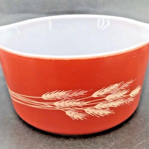 May include: A red Pyrex mixing bowl with a white interior and two handles. The exterior features a wheat stalk design in a cream color. This vintage bowl is ideal for kitchen use.
