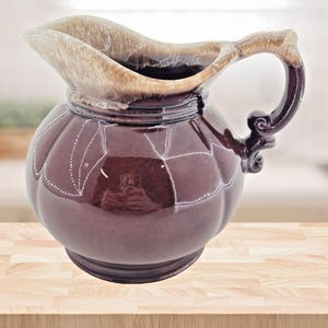 Puede incluir: Jarra decorativa, bulbosa, con cuerpo granate oscuro y borde y asa de color beige claro. La jarra tiene una forma única con una abertura ancha y un asa curvada. Líneas punteadas blancas acentúan el cuerpo.