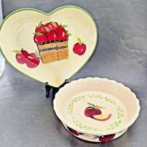 May include: Two ceramic serving dishes. One is heart-shaped with a green rim and a painted basket of red apples. The other is round with a scalloped edge and a painted apple and leaf design. Both dishes have a cream-colored background.