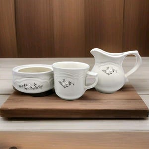 May include: A white ceramic creamer, sugar bowl, and cup with a floral design. The set is on a wooden tray.