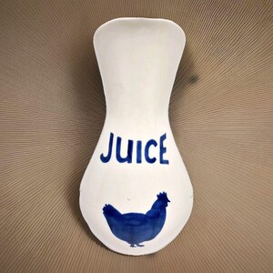 Könnte beinhalten: Weißer Keramik-Löffelhalter mit einer blauen Hühner-Silhouette und dem Wort "Juice" in blauen Buchstaben.