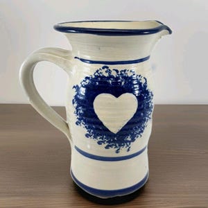 Puede incluir: Una jarra de cerámica con base blanca, adornada con detalles azules y un diseño de corazón. La jarra tiene un asa y un pico vertedor, perfecta para servir bebidas. El corazón está rodeado por un patrón azul en forma de corona.