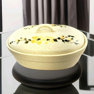 以下が含まれることがあります： クリーム色のセラミックキャセロール皿と蓋。 皿は斑点のある仕上げで、黄色の花の模様で飾られています。 蓋には小さなハンドルが付いています。 皿は黒い表面に置かれています。