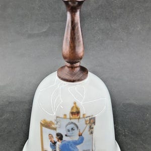 May include: A white ceramic bell with a wooden handle and clapper. The bell features a painting by Norman Rockwell, with the text "Norman Rockwell 1894-1978". The bell has a gold rim and a dark brown handle.