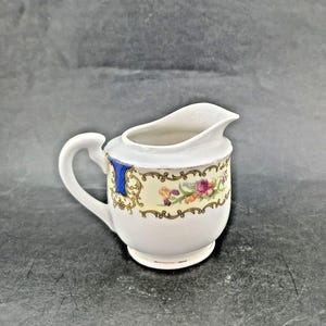 May include: A small, white ceramic creamer with a curved handle and spout. The creamer features a floral design with gold trim and a blue accent. The creamer is approximately 4 inches tall.