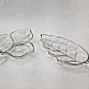 Puede incluir: Dos fuentes de vidrio transparente con forma de hoja. Una fuente tiene tres hojas conectadas, y la otra es una sola hoja alargada. Ambas tienen detalles de venas grabadas y están sobre un fondo blanco.