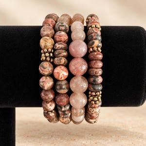 May include: A set of four beaded bracelets featuring a variety of natural stone beads in shades of brown, tan, pink, and gray. The bracelets include round and disc-shaped beads, accented with small metallic gold-toned spacer beads throughout.