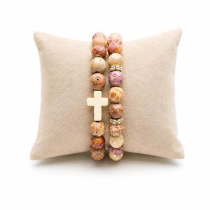 May include: Two beaded bracelets featuring round, multi-colored stone beads in shades of tan, brown, and mauve. One bracelet includes a central white cross charm, while the other features two small gold-toned spacers with embedded clear crystals.