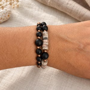 May include: A double-strand beaded bracelet featuring matte black spherical beads, cylindrical stone beads with a mottled grey and tan texture, and small metallic copper-toned spacer beads arranged in a repeating pattern.