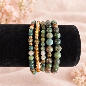 May include: A set of five beaded bracelets featuring various natural stone beads in shades of green, brown, and translucent amber. The bracelets include metallic gold-toned spacers with ribbed textures interspersed between the round beads.