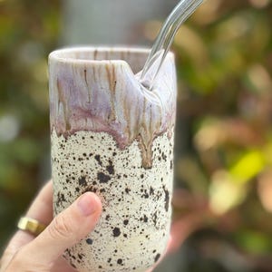May include: A handmade ceramic tumbler with a speckled cream base and a lavender and brown glazed top. A clear glass straw is inserted into the tumbler. The tumbler is held in a hand.