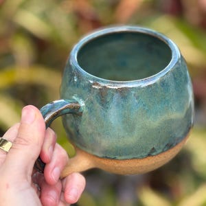 Könnte beinhalten: Handgefertigte Keramiktasse mit runder Form und türkisfarbener Glasur. Die Tasse hat einen braunen Boden und einen gebogenen Henkel. Das Innere der Tasse ist dunkler türkis. Die Tasse wird in einer Hand gehalten.