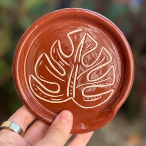 May include: A round, terracotta-colored ceramic spoon rest. The rest features a raised, white, stylized monstera leaf design in the center. The spoon rest is held in a hand, with a blurred background of green and red.
