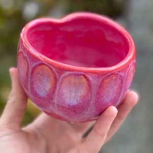 Puede incluir: Un cuenco de cerámica en forma de corazón en tonos rosa y rojo. El cuenco tiene un acabado brillante y un patrón texturizado en el exterior. El interior es de un rosa vibrante y sólido. El cuenco se sostiene en una mano sobre un fondo verde borroso.