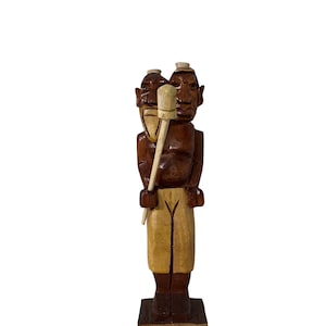 May include: A carved wooden statue depicting two figures with a shared face and a light-colored wooden tool. The statue is primarily brown with tan and light brown accents, standing on a dark brown base.