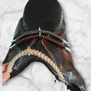 May include: A brown leather saddle with a tan braided rope attached to a metal ring. The saddle is on a white marble surface.
