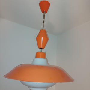 May include: A mid-century modern pendant light fixture with a white and orange color scheme. The light has a large, saucer-shaped shade with a smaller, cone-shaped shade above it.