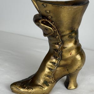May include: A vintage-style, bronze-colored boot-shaped vase. The boot features a high heel, button details, and a decorative buckle. The vase has a textured surface and a flared top, evoking a classic, antique aesthetic. The boot measures approximately 7 inches in height.