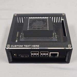 May include: A black computer case with a clear top, featuring the text "CUSTOM TEXT HERE." The case has a rectangular shape with multiple ports and a cooling system on top. The design is modern and sleek.