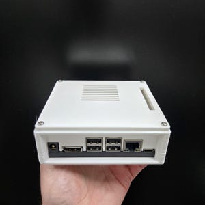 May include: A white rectangular electronic device with multiple ports and a vent on top. The device has a power port, HDMI port, USB ports, an Ethernet port, and a USB-C port. The device is held in a hand against a black background.