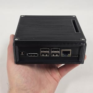 May include: Black rectangular electronic device with a ridged top, held in a hand. The device has multiple ports, including USB, HDMI, and Ethernet. The case is black and has a small opening on the side.