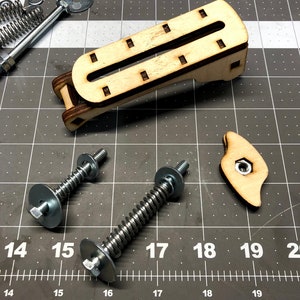 May include: Two silver metal screws with a spring and a washer on each end. The screws are on a gray grid surface with a ruler marked in inches. A wooden piece with a slot in the middle is also on the surface.