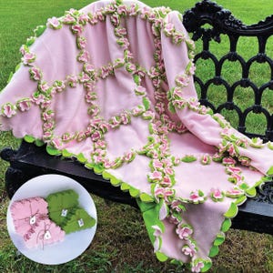 May include: A light pink fleece blanket featuring a grid of three-dimensional pink and green fabric flowers sewn along the seams. A circular inset shows three stacks of fabric flower cutouts labeled A, B, and C with twine ties.
