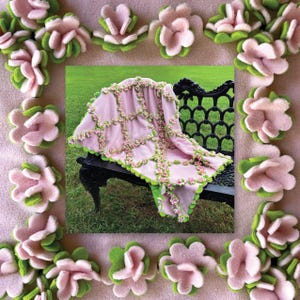 Könnte beinhalten: Eine rosa Decke mit grünen Verzierungen und floralen Akzenten liegt über einer schwarzen Bank. Die Decke ist von einem Rand aus rosa und grünen Filzblumen umgeben, wodurch ein dekoratives und skurriles Design entsteht.