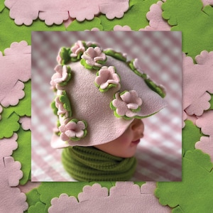 May include: A light pink hat adorned with pink and green felt flowers. The hat is paired with a green scarf. The background features pink and green felt cutouts, creating a whimsical, floral theme.