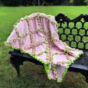 May include: A pink fleece blanket with green and pink floral accents. The blanket is draped over a black wrought iron bench. The edges and seams are decorated with green and pink flowers, creating a decorative and cozy look.