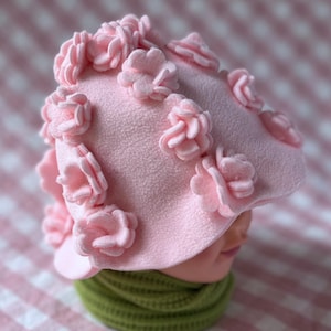 May include: A light pink hat adorned with multiple pink fabric flowers. The hat has a rounded crown and a small brim. A green scarf is visible below the hat. The hat is displayed on a mannequin head.