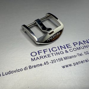 May include: A polished silver watch buckle with the brand name "PANERAI" engraved on the curved surface. The buckle is rectangular with a central bar and two side bars.