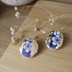 Può includere: Un paio di orecchini in ceramica blu e bianca con dettagli dorati. Gli orecchini presentano un design floreale con un fiore bianco e petali blu.