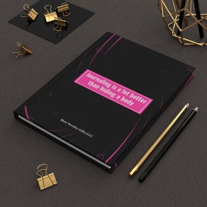 May include: Black journal with pink abstract design and the text "Journaling is a lot better than hiding a body". The journal is on a gray surface with gold pencils and paper clips.