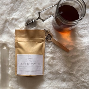 May include: A brown paper bag with a label that reads "Organic & Wildcrafted Tea Hand-Made in Small Batches Divine Energy Tea" with a white background. The bag is sitting on a white fluffy surface with a glass jar of tea, a tea strainer, and a wooden spoon.