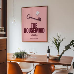 May include: A framed poster with a pink background features a key and a tooth graphic. The text "THE HOUSEMAID" is displayed in large, dark red letters. Below, smaller text reads "IN THEATRES DECEMBER 19" and a quote. The poster is in a wooden frame.