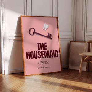 May include: A framed movie poster for "The Housemaid" with a pink background. The poster features a key and a tooth graphic. The title is in large, dark red letters. The poster is leaning against a white wall.