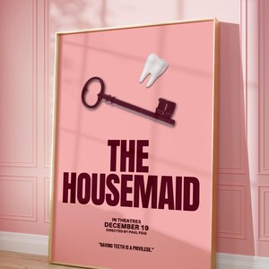 May include: A framed movie poster for "The Housemaid" with a pink background. The poster features a key and a tooth graphic. The movie title is in large, dark red letters. The release date is December 19.