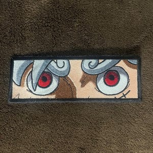 Peut inclure: Gros plan sur un patch brodé blanc et gris avec deux yeux rouges. Les yeux sont entourés de volutes blanches et grises. Le patch est encadré d'une bordure noire.