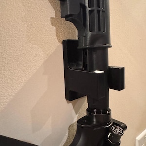 May include: Black firearm mounted on a wall. The firearm is held in place by a black bracket. The image shows the rear of the firearm, including the stock and the lower receiver.