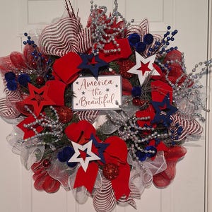 Puede incluir: Una corona patriótica roja, blanca y azul con un letrero que dice "America the Beautiful". La corona está decorada con estrellas, lazos y brillo.